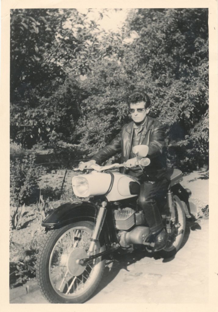 Eine Person sitzt auf einenm Motorrad. Die Person trägt Lederkleidung und einen Sonnenbrille und hat kurze, gelockte dunkle Haare. Im Hintergrund sind Bäume. Das Bild ist in schwarz/weiß.